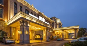 Illuminated grand exterior of a luxury hotel at dusk, featuring a columned entrance with yellow lights and parked cars in front.